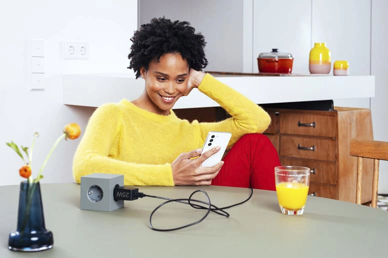 Een jonge vrouw zit met een Samsung snellader aan tafel en glimlacht in haar smartphone.