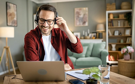 Een man werkt thuis in zijn woonkamer op zijn laptop en telefoneert met een headset.