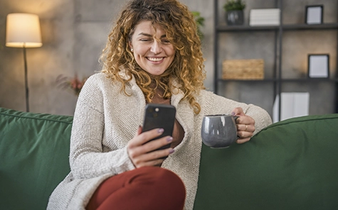 Een vrouw zit op een gezellige groene bank en gebruikt haar smartphone om haar slimme huis te bedienen.