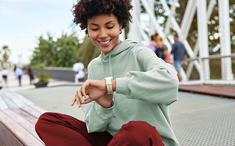 Een jonge vrouw in een groene trui en rode broek zit buiten op een muurtje en kijkt goedgehumeurd op haar smartwatch