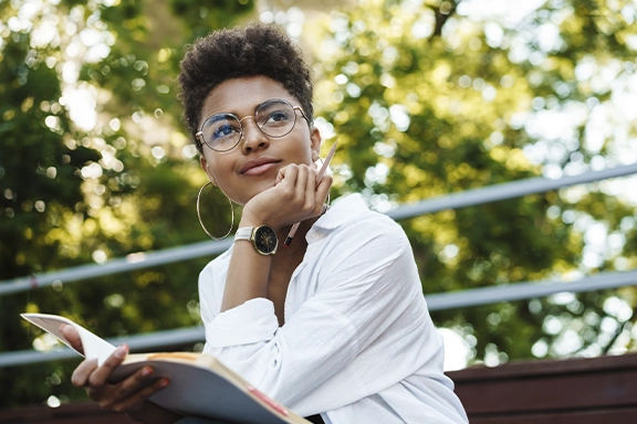 Een vrouw in een zakelijke look zit buiten op een bankje onder bomen en staart dromerig langs de camera. Ze draagt de Hama Smartwatch “7000” om haar pols.