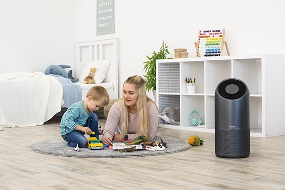Een jonge vrouw ligt met een kind op de grond in de kinderkamer en speelt. Daarnaast staat de Hama "Smart" luchtreiniger